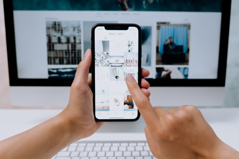 Hands holding a smartphone while browsing a social media app in front of a desktop computer